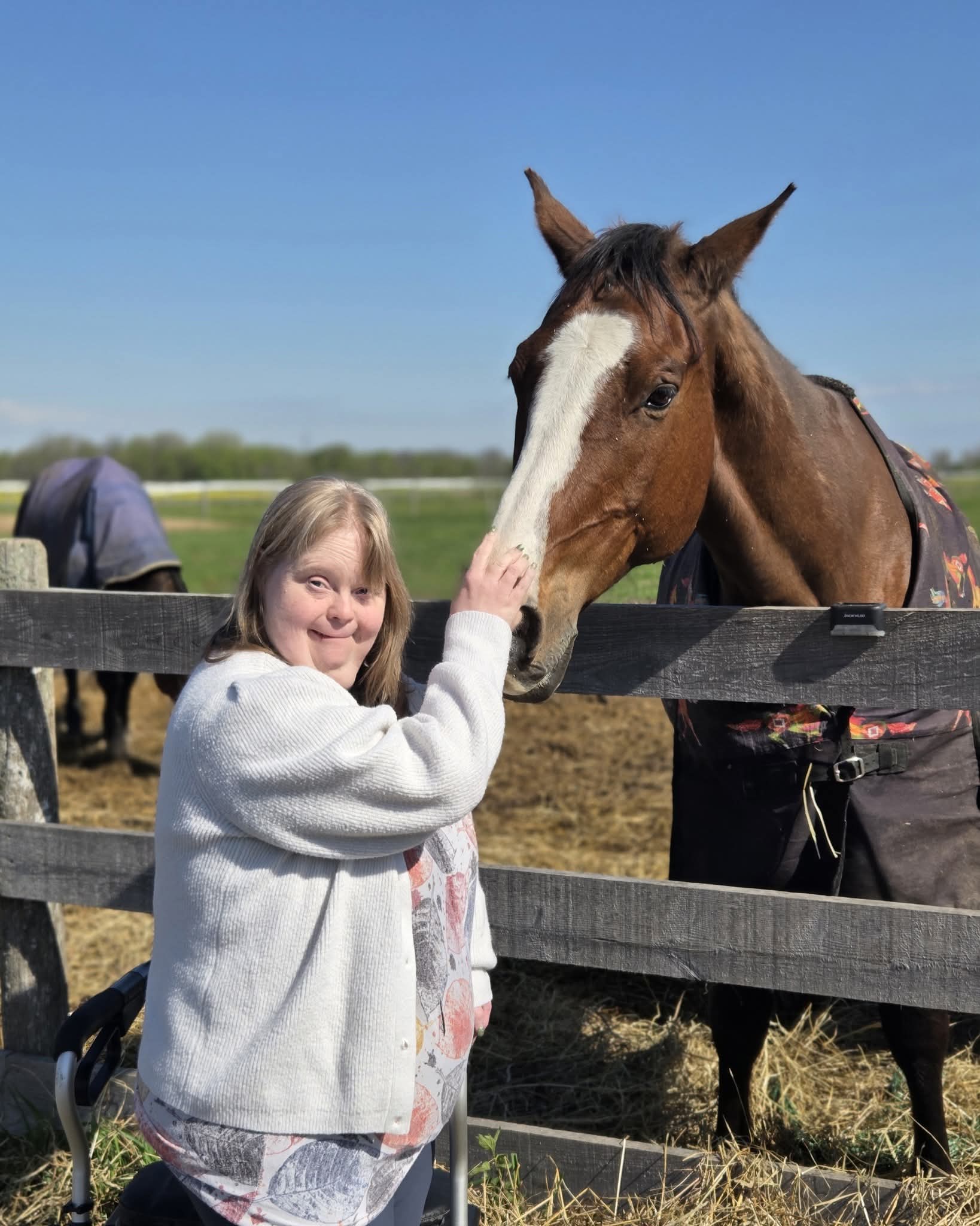 A smiling participant stands with her walker and pets a horse over a wooden paddock fence under a blue sky